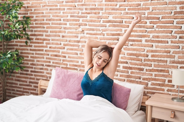 Young blonde woman waking up stretching arms at bedroom