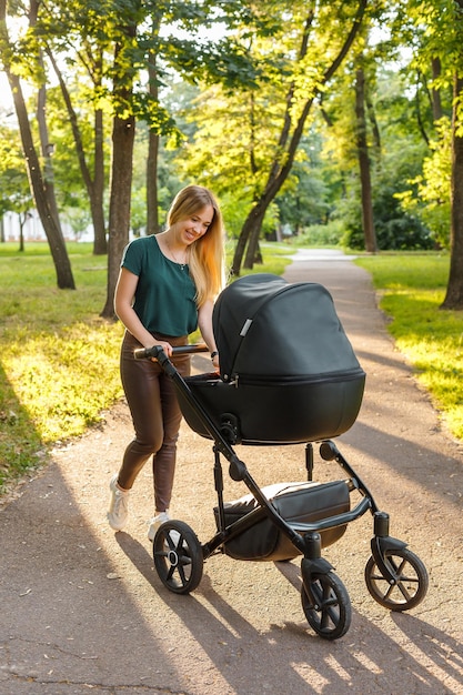 Young blonde woman walking with black stroller in summer park happy mother with baby in pram