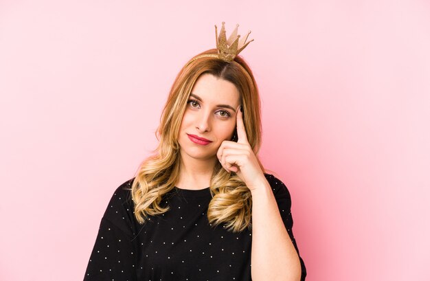 Photo young blonde woman wearing a crown pointing temple with finger, thinking, focused on a task.