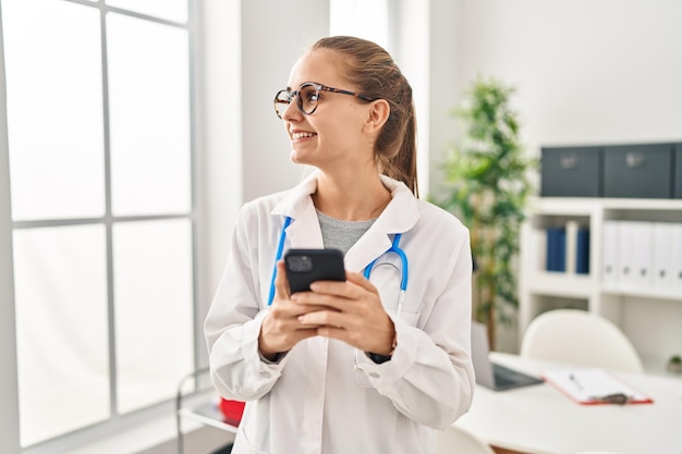 Young blonde woman wearing doctor uniform using smartphone working at clinic