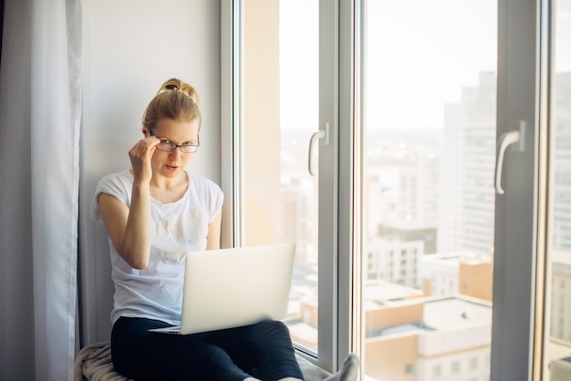 Young blonde woman wearing glasses in white t-shirt working on laptop at home, sitting on the windowsill. Freelancer and remote business. Window background. Copy space.