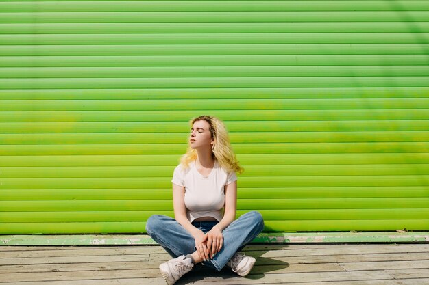 Young blonde woman wearing a white top and blue jeans sitting on a wooden floor on a vivid green wall