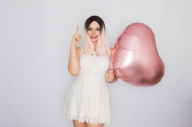 Young blonde woman in white lace dress holding big pink heart balloon in her hands over white background