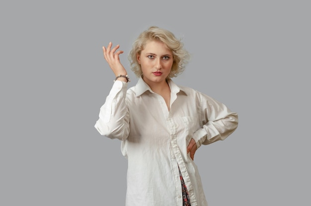 Young blonde woman in a white shirt looks at the camera hand on hand other hand raised up Surprised