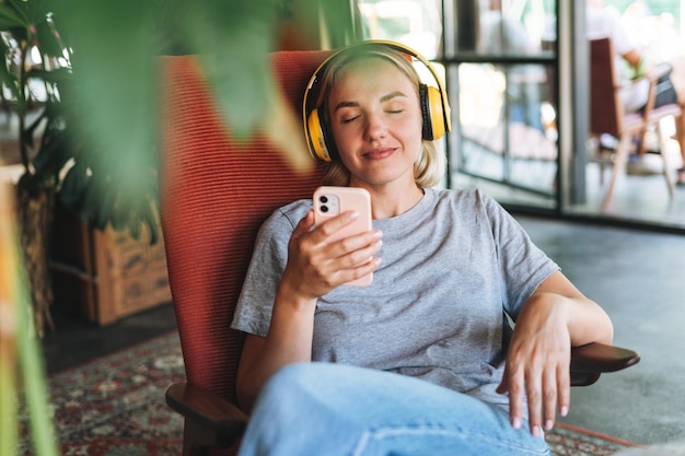 Young blonde woman with close eyes in yellow headphones enjoys music with mobile phone at cafe