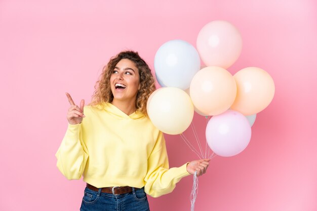 Young blonde woman with curly hair catching many balloons isolated on pink wall pointing with the index finger a great idea