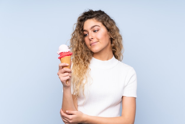 Young blonde woman with curly hair holding a cornet ice cream on blue wall with sad expression