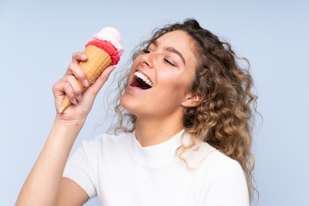 Young blonde woman with curly hair holding a cornet ice cream on blue wall