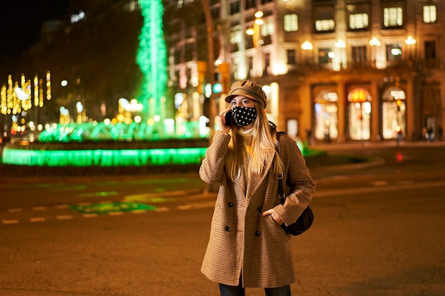 Young blonde woman with mask talking on the phone outdoors at night. In the background there are many city lights. Winter atmosphere.