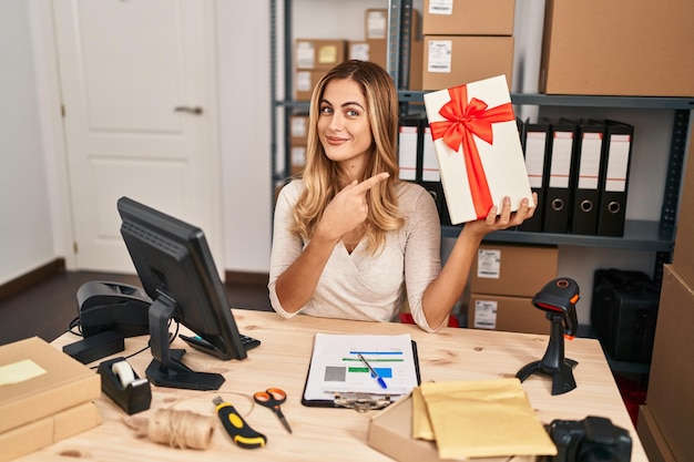 Young blonde woman working at small business ecommerce holding gift smiling happy pointing with hand and finger