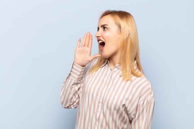 Young blonde woman yelling loudly and angrily to copy space on the side, with hand next to mouth