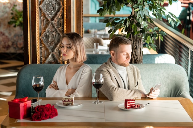 Young bored or offended woman sitting by served table in restaurant with her boyfriend scrolling in smartphone near by