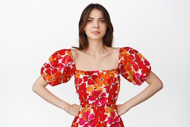 Young bothered girl in red dress holding hands on waist frowning and looking disappointed feeling doubtful or concerned standing against white background