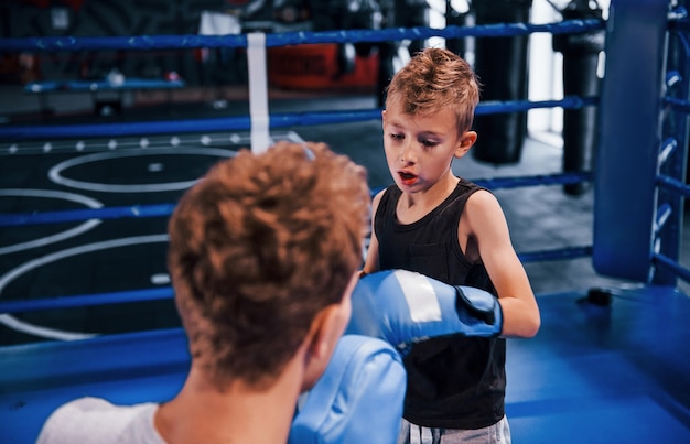 Young boxing coach is helping little boy in protective wear on the ring between the rounds.