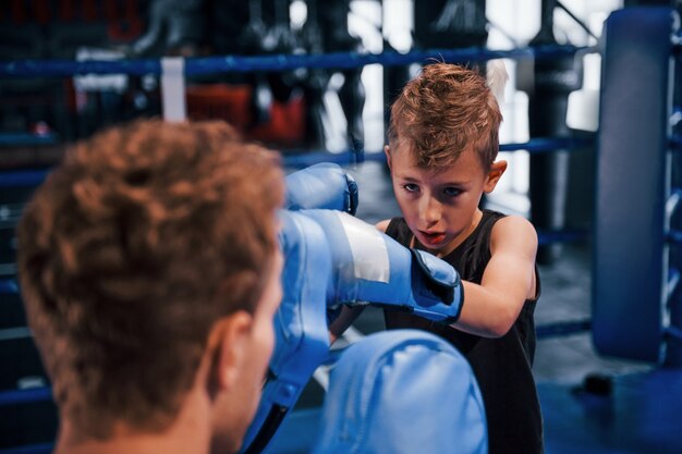 Young boxing coach is helping little boy in protective wear on the ring between the rounds.