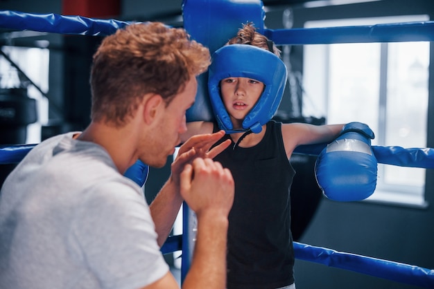 Photo young boxing coach is helping little boy in protective wear on the ring between the rounds.