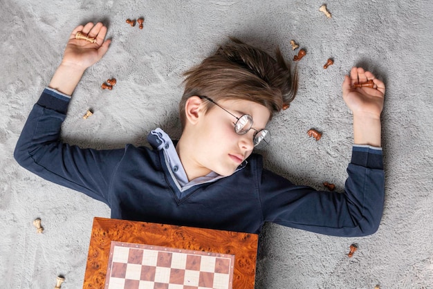 Young boy chess winner in glasses lying on the carpet around chess pieces and checkmate, top view