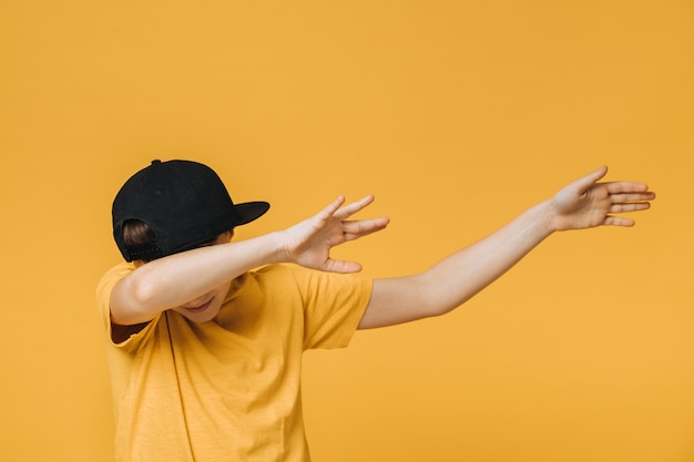 Young boy dressed in yellow t-shirt and black baseball cap over yellow background makes teenagers dab gesture, moves his hands aside, covering his face, that means i did it. Youth culture concept.