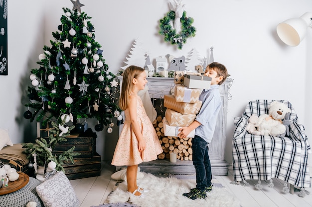 Young boy presenting gift boxes to pretty little girl in room with cristmas tree and fireplace. Winter holidays.