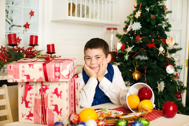 young boy sitting at the table with presents near and the christmas tree