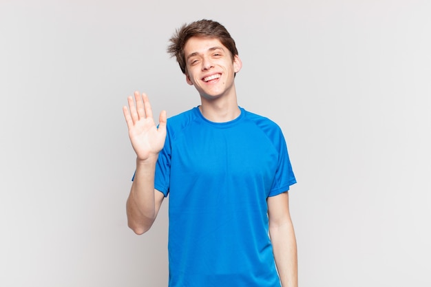 young boy smiling happily and cheerfully, waving hand, welcoming and greeting you, or saying goodbye