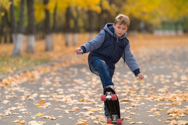 Young boy in warm jacket doing skateboard trick viewed from his front in full length on park walkway covered with yellow leaves and trees