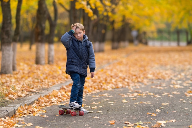 Young boy in warm jacket with skateboard holding his hand to forehead, as if it is hurt, standing alone in the park. Yellow leaves and trees