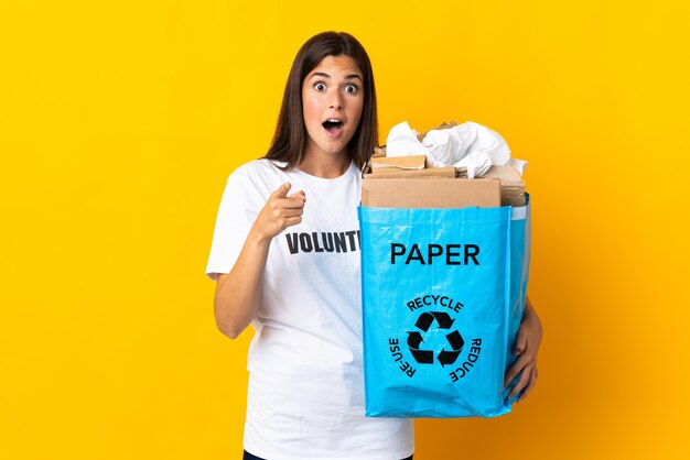 Young brazilian girl holding a recycling bag full of paper to recycle isolated on yellow background surprised and pointing front