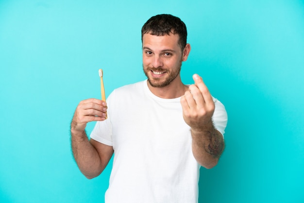 Young Brazilian man brushing teeth isolated on blue background making money gesture