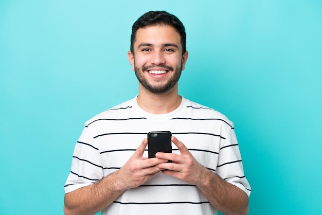 Young Brazilian man isolated on blue background looking at the camera and smiling while using the mobile