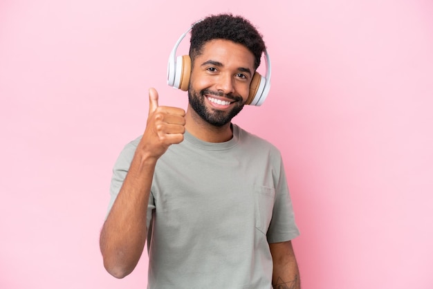 Young Brazilian man isolated on pink background listening music and with thumb up