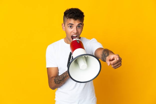 Young Brazilian man over isolated wall shouting through a megaphone to announce something while pointing to the front