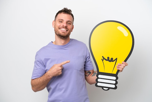 Young Brazilian man isolated on white background holding a bulb icon and pointing it