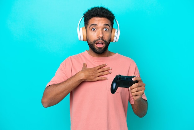 Young brazilian man playing with a video game controller isolated on blue background surprised and shocked while looking right