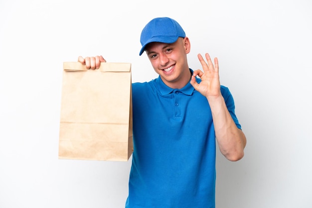 Young Brazilian man taking a bag of takeaway food isolated on white background showing ok sign with fingers