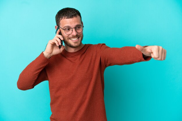 Young Brazilian man using mobile phone isolated on blue background giving a thumbs up gesture