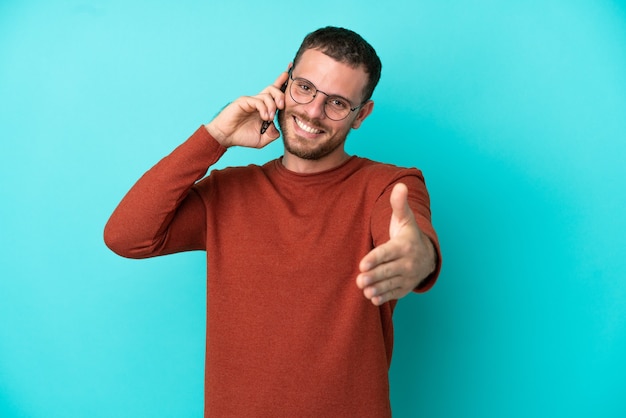 Young Brazilian man using mobile phone isolated on blue background shaking hands for closing a good deal