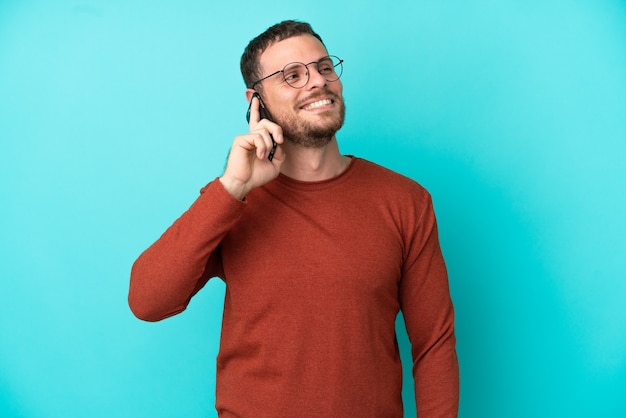 Young Brazilian man using mobile phone isolated on blue background thinking an idea while looking up