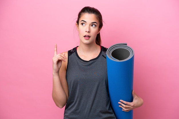 Young Brazilian sport woman going to yoga classes while holding a mat isolated on pink background thinking an idea pointing the finger up