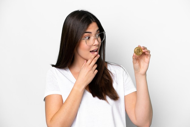 Young Brazilian woman holding a Bitcoin isolated on white background with surprise and shocked facial expression