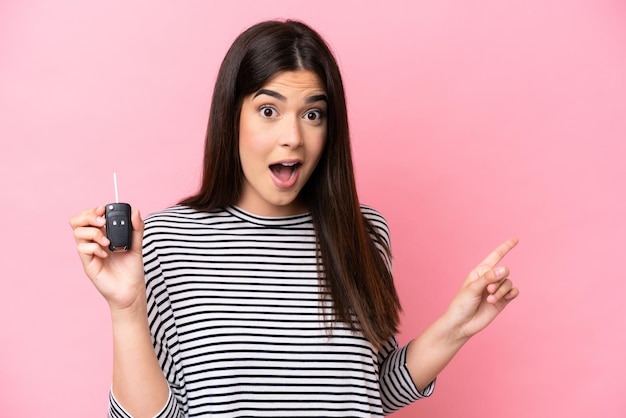 Young Brazilian woman holding car keys isolated on pink background surprised and pointing finger to the side