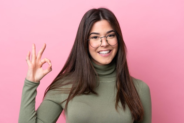 Young Brazilian woman isolated on pink background With glasses and doing OK sign