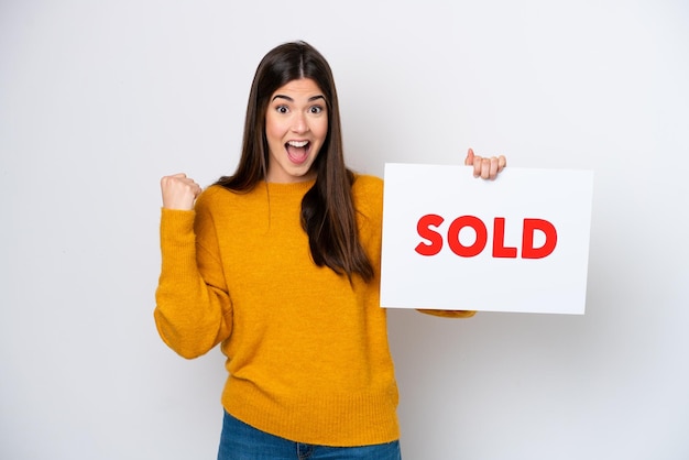 Young Brazilian woman isolated on white background holding a placard with text SOLD and celebrating a victory