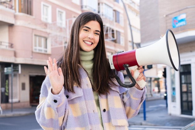 Young Brazilian woman at outdoors holding a megaphone and saluting with hand with happy expression