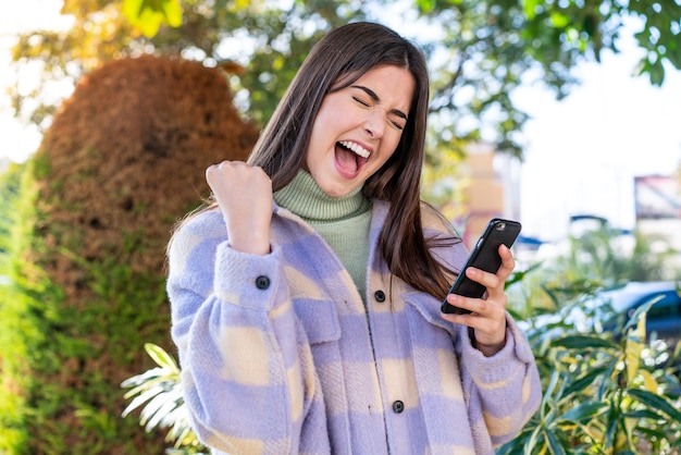 Young Brazilian woman in a park with phone in victory position