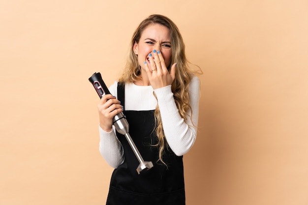 Young brazilian woman using hand blender isolated on beige happy and smiling covering mouth with hand