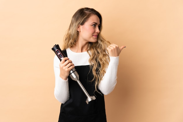 Young brazilian woman using hand blender isolated on beige  pointing to the side to present a product