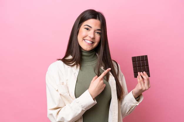 Young Brazilian woman with chocolat isolated on pink background pointing to the side to present a product