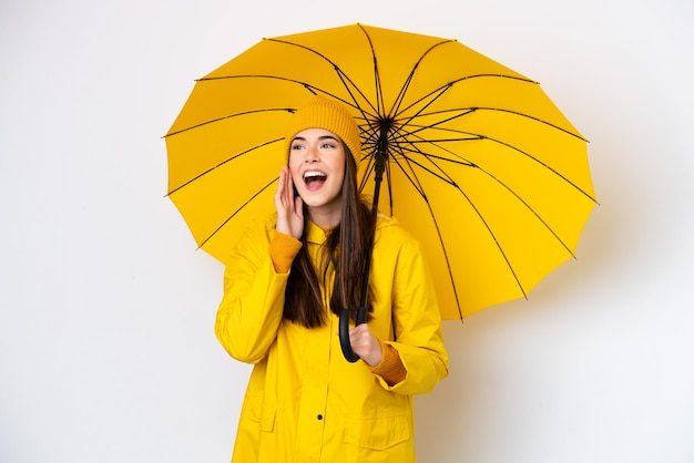 Young Brazilian woman with rainproof coat and umbrella isolated on white background shouting with mouth wide open to the side