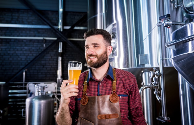 Young brewer wearing a leather apron is testing beer at a modern brewery